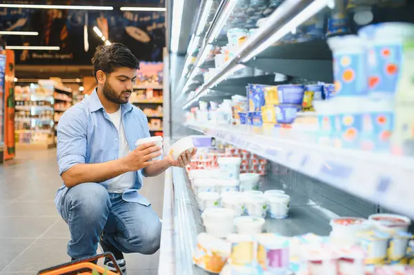 Genç adam yoğurtları karşılaştırırken bir marketin soğutulmuş süt koridorunda çömeliyor.