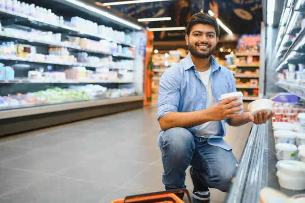 Gülümseyen bir müşteri marketteki soğutulmuş rafın yanında çömelmiş iki yoğurt konteynırını karşılaştırıyor.