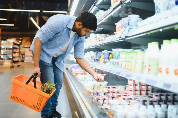 Müşteri, modern bir süpermarketin soğutuculu koridorundan çeşitli süt ürünleri seçiyor. Turuncu sepeti sağlıklı seçeneklerle dolduruyor.