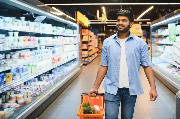 Genç Hintli bir adam süpermarket koridorlarında yürürken bir alışveriş sepeti taşıyor, içinde bakkaliye bulunan soğutulmuş raflara bakıyor.
