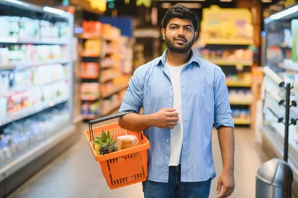 Parlak ve düzenli bir süpermarkette alışveriş yaparken alışveriş yaparken alışveriş sepetini taşıyan genç Hintli bir adam perakende deneyiminin tadını çıkarıyor.