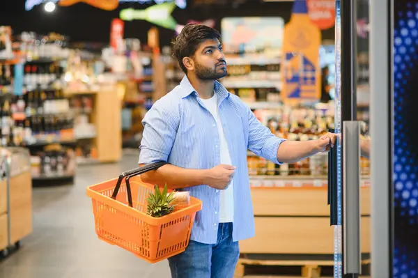 Genç Hintli bir adam, açık ve düzenli bir market koridorunda buzdolabının kapısını açarken elinde alışveriş sepeti taşıyor.