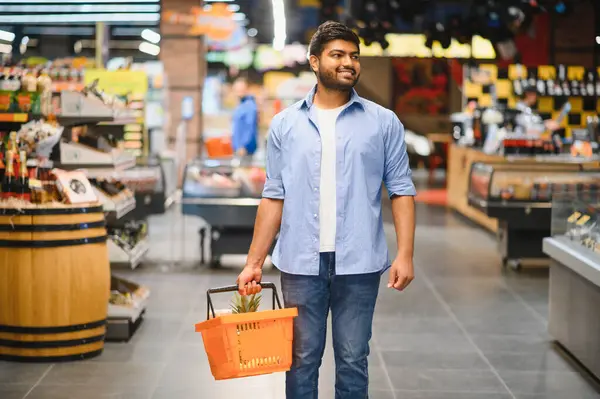 Genç Hintli bir adam bir alışveriş sepeti taşıyor ve bir market koridorunda yürüyor, alışveriş deneyiminin tadını çıkarıyor.