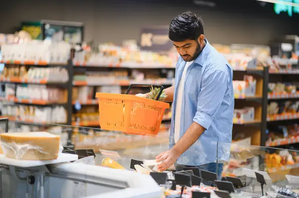 Bir markette elinde turuncu bir alışveriş sepetiyle soğutucudan peynir seçen odaklanmış bir müşteri.