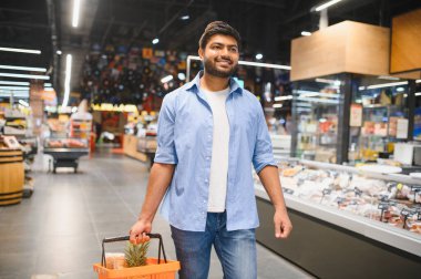 Bir süpermarket koridorunda yürürken alışveriş deneyiminin tadını çıkaran bir alışveriş sepeti taşıyan genç Hintli.