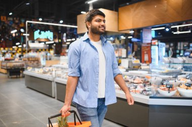 Gülümseyen Hintli müşteri market koridorlarında yürürken sepet taşıyor, taze ürünler seçiyor.