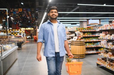 Genç Hintli müşteri bir alışveriş sepeti taşıyor, taze ürün ve bakkaliyelerle dolu, parlak ve modern bir süpermarket koridorunda yürüyor.