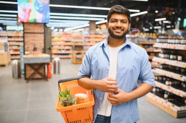 Genç Hintli adam elinde alışveriş sepeti tutarken gülümsüyor. İçi alışveriş dolu. Canlı bir süpermarket koridorunda eğleniyor.