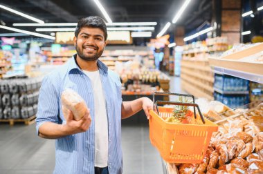 Genç Hintli adam elinde ananasla ekmek ve alışveriş sepeti tutuyor, süpermarketten alışveriş yaparken gülümsüyor.