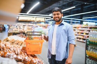 Genç Hintli bir adam market alışverişi yapıyor, içinde ananas olan turuncu bir alışveriş sepeti tutuyor, parlak ışıklı bir süpermarkette.
