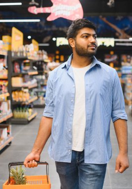 Müşteri bir alışveriş sepeti taşıyor, bakkal koridorunda yürüyor, rafları taze yiyecek ve ürünlerle dolduruyor.