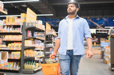Markette yürürken alışveriş sepeti taşıyan genç Hintli adam raflardan ürünler seçiyor.