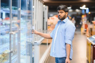 Odaklanmış Hintli adam soğutuculu bölüm kapısını açıyor. Parlak ışıklı bir süpermarkette market alışverişi yapıyor.