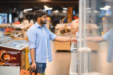Market alışverişi yapan genç Hintli adam, yeni seçeneklerle dolu, parlak, modern bir süpermarkette soğutulmuş bir koridorun kapısını açıyor.