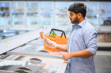 Plastik eldiven giyen bir müşteri marketteki dondurulmuş eti seçiyor, elinde ananasla alışveriş sepeti tutuyor.