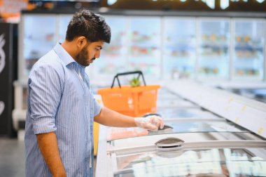 Plastik eldiven giyen genç Hintli adam süpermarketteki dondurulmuş gıda ürünlerini seçiyor.