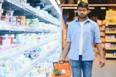 Genç Hintli bir adam markette dondurulmuş süt ürünleri reyonunda yürürken alışveriş sepsiyonu taşıyor.