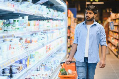 Genç Hintli adam marketin buzdolabı bölümünde yürüyor, alışveriş sepeti taşıyor ve sebze seçiyor.