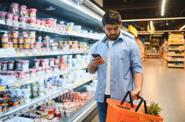 Akıllı telefon kullanan ve marketten alışveriş sepeti tutan konsantre Hintli genç bir adam.