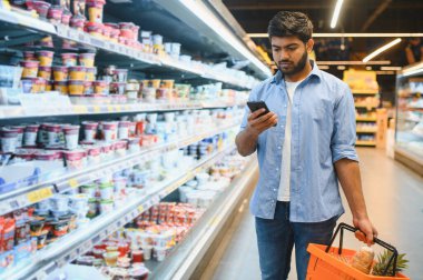 Portakal rengi bir alışveriş sepeti taşırken akıllı telefon kullanan genç bir adam, yoğun bir süpermarketin koridorlarında geziniyor.