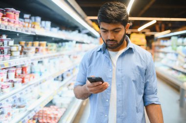 Markette alışveriş yaparken akıllı telefon kullanan, alışveriş listesini kontrol eden ya da fiyatları karşılaştıran Hintli adam.