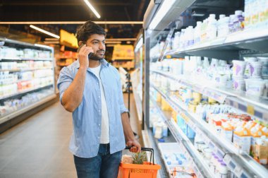 Elinde alışveriş sepeti olan genç bir Hintli süpermarkette süt ürünlerini seçerken telefonla konuşuyor.