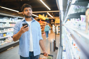 Genç adam alışveriş listesini akıllı telefondan kontrol ederken marketin kalabalık koridorunda yiyecek seçiyor.
