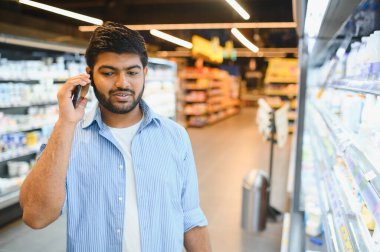 Genç Hintli bir adam akıllı telefon kullanırken markette taze yiyecekler seçiyor ve modern bir alışveriş deneyiminin tadını çıkarıyor.