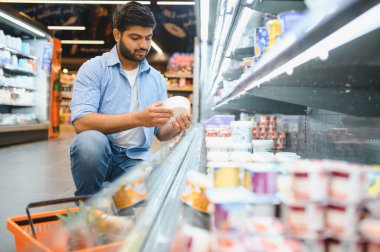 Genç Hintli bir adam süpermarketin buzdolabında çömelmiş ürünlerin malzemelerini kontrol ediyor.
