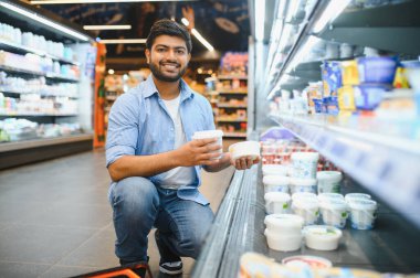Gülümseyen bir müşteri markette soğutulmuş rafların yanında çömelirken iki yoğurt kabını karşılaştırıyor.