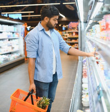 Market alışverişine odaklanmış Hintli bir adam, elinde bir sepet tutuyor ve modern bir süpermarkette soğutulmuş bir vitrinden süt ürünleri seçiyor.
