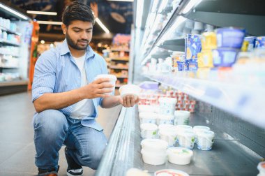 Genç Hintli bir adam yoğurt markalarını karşılaştırırken modern bir marketin dondurulmuş süt ürünleri reyonunda çömeliyor.