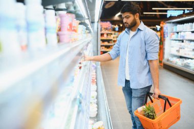 Soğutulmuş raflardan süt ürünlerini seçen odaklanmış genç Hintli bir adam, içinde ananas olan alışveriş sepetini modern bir süpermarkette tutuyor.