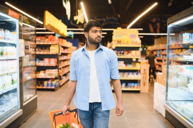 Genç Hintli bir adam bir alışveriş sepeti taşıyor, market koridorunda yürüyor, taze ürünler seçiyor ve alışveriş deneyiminin tadını çıkarıyor.