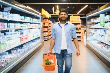 Parlak bir süpermarket koridorunda yürüyen gülümseyen Hintli bir adam, içi taze bakkaliye ve sağlıklı ürünlerle dolu bir alışveriş sepeti taşıyor.