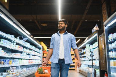 Genç Hintli adam süpermarket koridorunda elinde birkaç eşyayla yürüyor, gülümsüyor ve etrafa bakıyor.
