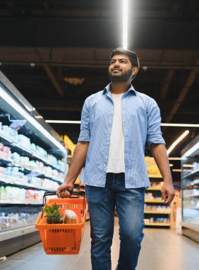 Müşteri, canlı bir süpermarkette alışveriş yaparken içi yiyecek dolu bir sepet taşıyor. Organize koridorlarda ve raflarda geziniyor.