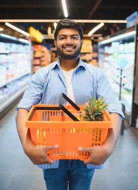 Marketten alışveriş yaparken ananaslı turuncu bir alışveriş sepeti taşıyan mutlu Hintli müşteri.