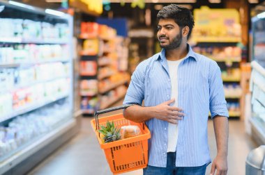 Markette yürürken ve ürünleri seçerken elinde alışveriş sepeti taşıyan genç Hintli bir adam.