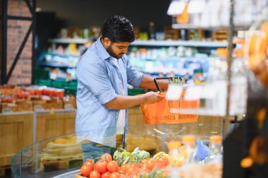 Elinde alışveriş sepeti olan genç Hintli bir adam modern bir süpermarketin soğutulmuş bölümünden taze sebze seçiyor.