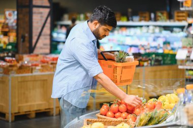 Genç Hintli adam taze ürün almak için alışveriş yapıyor, parlak ışıklı bir markette domates topluyor.