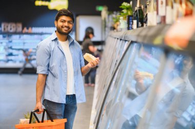 Hintli müşteri elinde dondurulmuş yiyeceklerle dolu bir alışveriş sepeti, canlı bir süpermarket ortamında market koridorlarında geziniyor.
