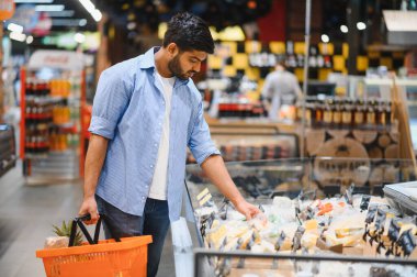 Müşteri marketin soğutulmuş bölümünden peynir seçiyor, elinde ekmek ve ananas ile alışveriş sepeti tutuyor.