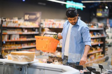 Modern bir süpermarkette soğutulmuş bölümden peynir seçen Hintli genç bir müşteri, elinde bir alışveriş sepeti tutuyor.