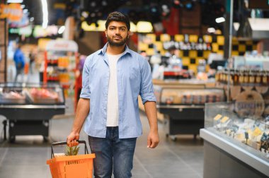 Genç Hintli bir adam market koridorlarında yürürken bir alışveriş sepeti taşıyor, taze ürünler ve bakkaliye seçiyor.