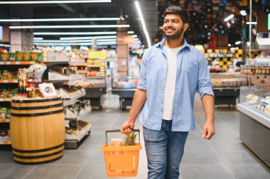 Genç Hintli adam markette yürürken alışveriş sepeti taşıyor, gülümsüyor ve başka tarafa bakıyor.