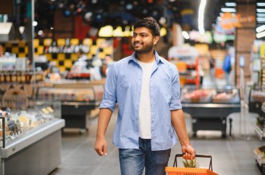 Bir süpermarket koridorunda yürürken elinde alışveriş sepeti taşıyan genç Hintli adam gülümsüyor ve etrafa bakıyor.