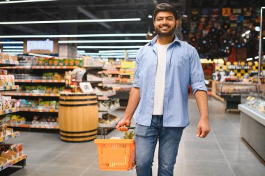 Market koridorlarında yürürken turuncu bir alışveriş sepeti taşıyan genç Hintli müşteri.