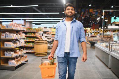 Müşteri, taze ürünler ve çeşitli malzemelerle çevrili, parlak, düzenli bir süpermarkette alışveriş yaparken bir sepet taşıyor.