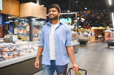 Markette elinde alışveriş sepetiyle dolaşan genç Hintli adam süpermarketten ürünler seçiyor.
